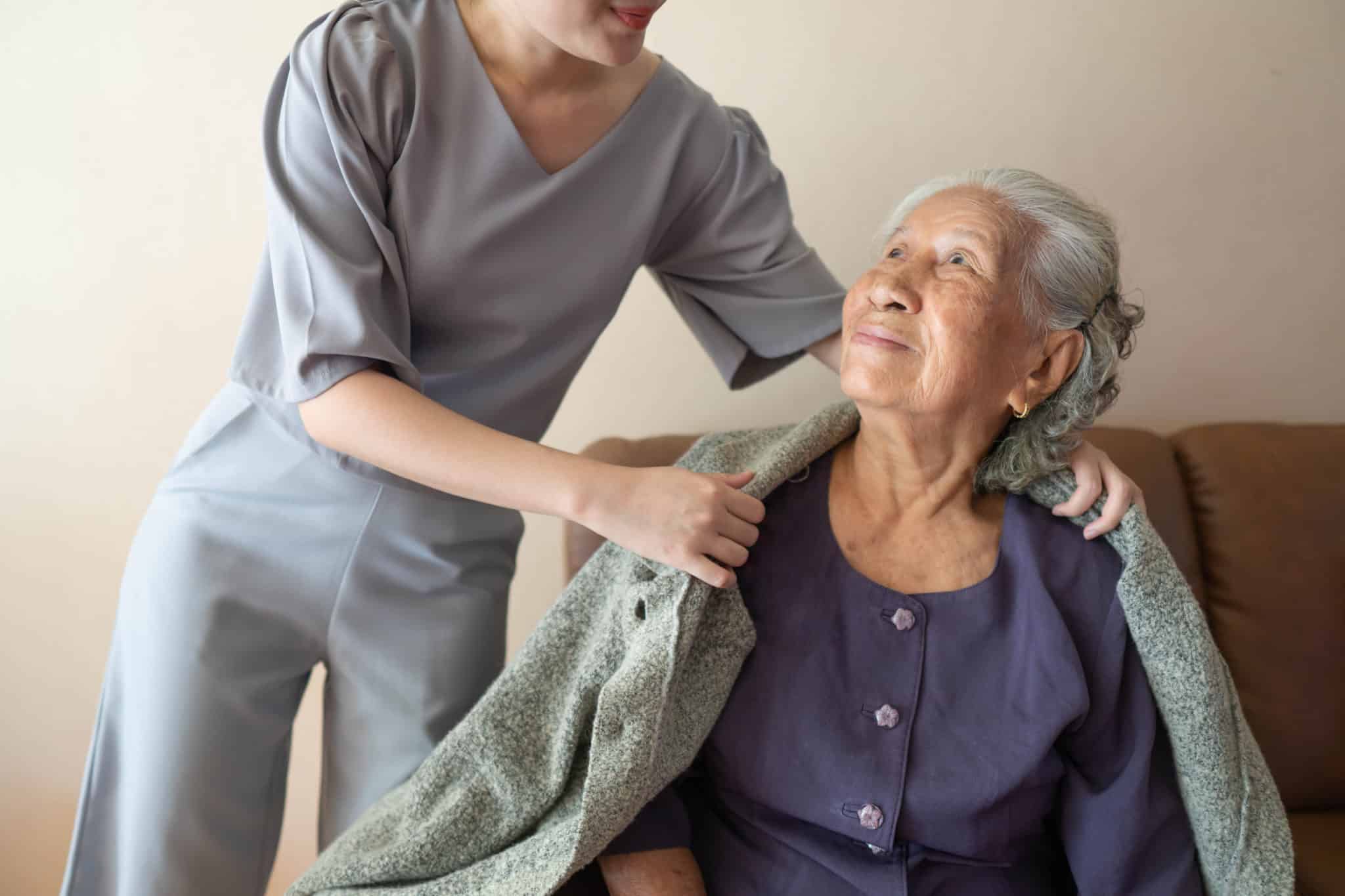 Caregiver covering senior woman with sweater at home. Happy elderly patient with friendly female nurse spending time together.