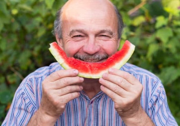 Mature caucasian man with mustache eating juicy water melon with pleasure and smiling. Healthy summer food