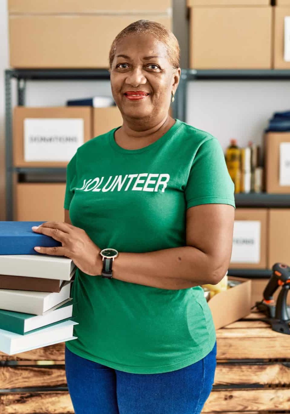 Senior African American Woman Wearing Volunteer Uniform Holding Books At Charity Center